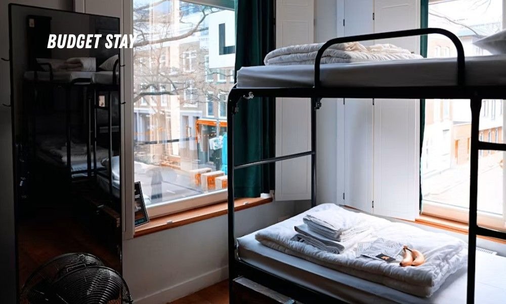 A clean hostel room with black metal bunk beds, bright sunlight streaming through a window, and a graphic that reads BUDGET STAY.