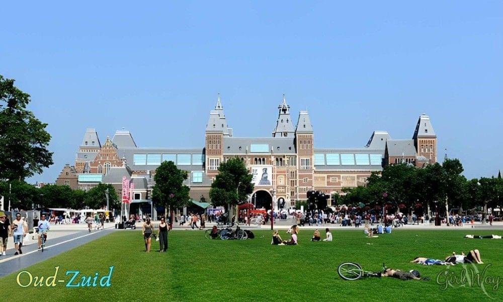 Oud-Zuid in Amsterdam with Rijksmuseum buildings rising above a green lawn under a clear blue sky near Museumplein and the Museum Quarter.